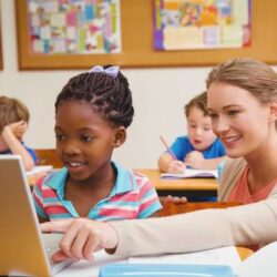 Female Teacher Showing Girl Laptop Screen Sitting At Table Smiling